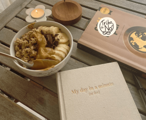 Mesa de madeira com uma taça de aveia, banana e manteiga de amendoim, uma vela acesa num suporte em forma de flor, um caderno de capa de linho e o logótipo da Tribo do Nó sobre um livro.